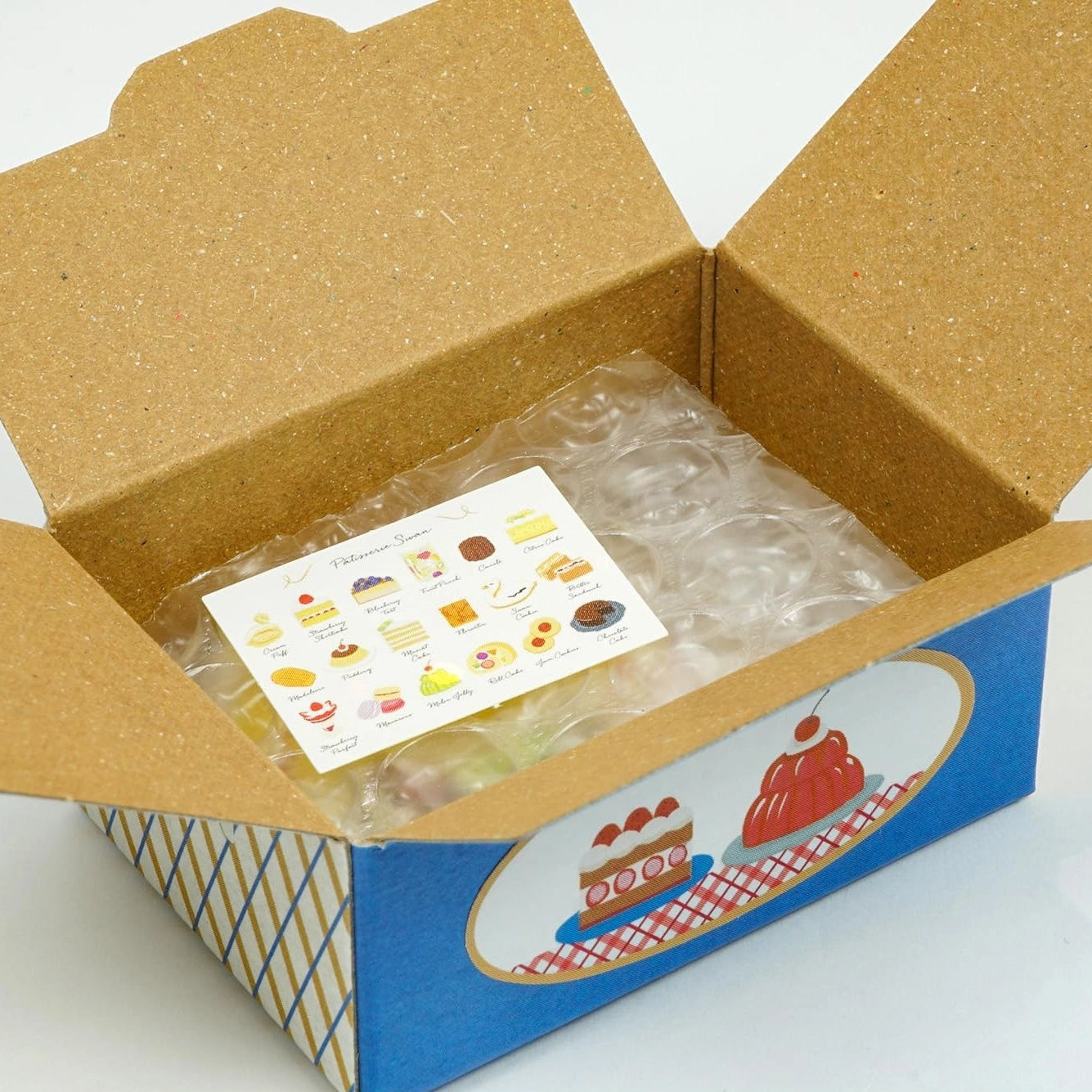 Open cardboard box with a blue base and clear plastic cover containing a card with illustrations.