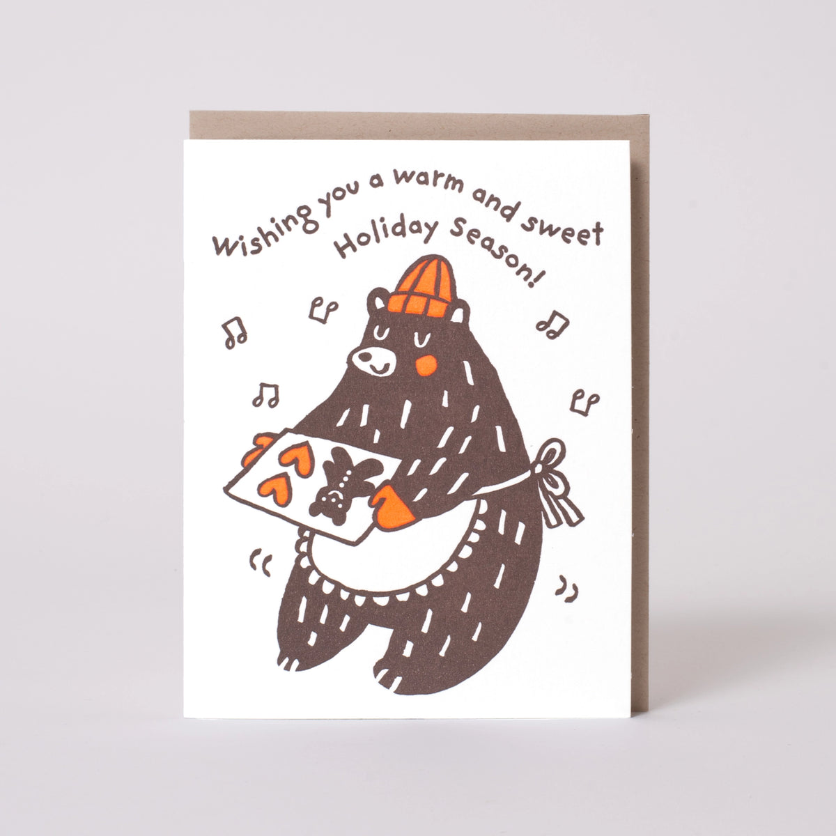 A white greeting card with a brown bear illustration holding a carrot, with the text 'Wishing you a warm and sweet Holiday Season!' above it. A brown envelope is partially visible behind the card against a light gray background.