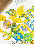 Colorful banana-themed stickers on a white surface with a cardboard box in the background.