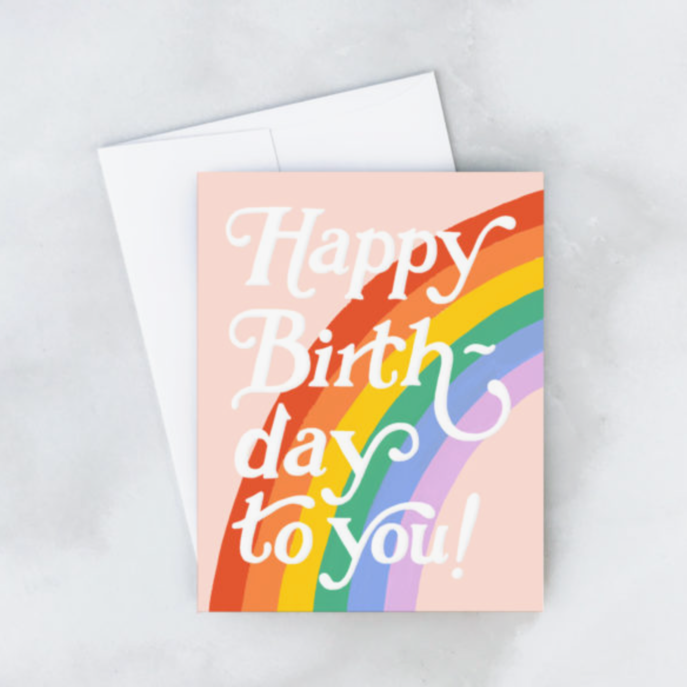 A bright, multicolored birthday card with the text 'HAPPY BIRTHDAY TO YOU!' and a rainbow design, placed on a marble surface with a matching envelope.
