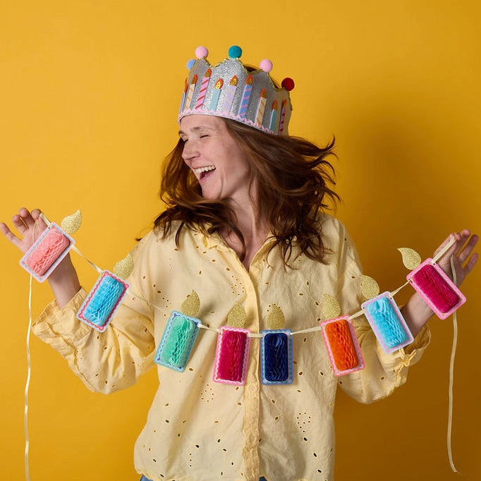 Woman wearing a colorful birthday crown and holding a banner with festive decorations against a yellow background