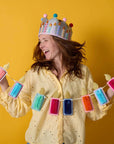 Woman wearing a colorful birthday crown and holding a banner with festive decorations against a yellow background