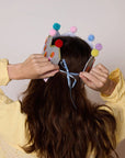 Person wearing a colorful headband with pom-poms against a plain background