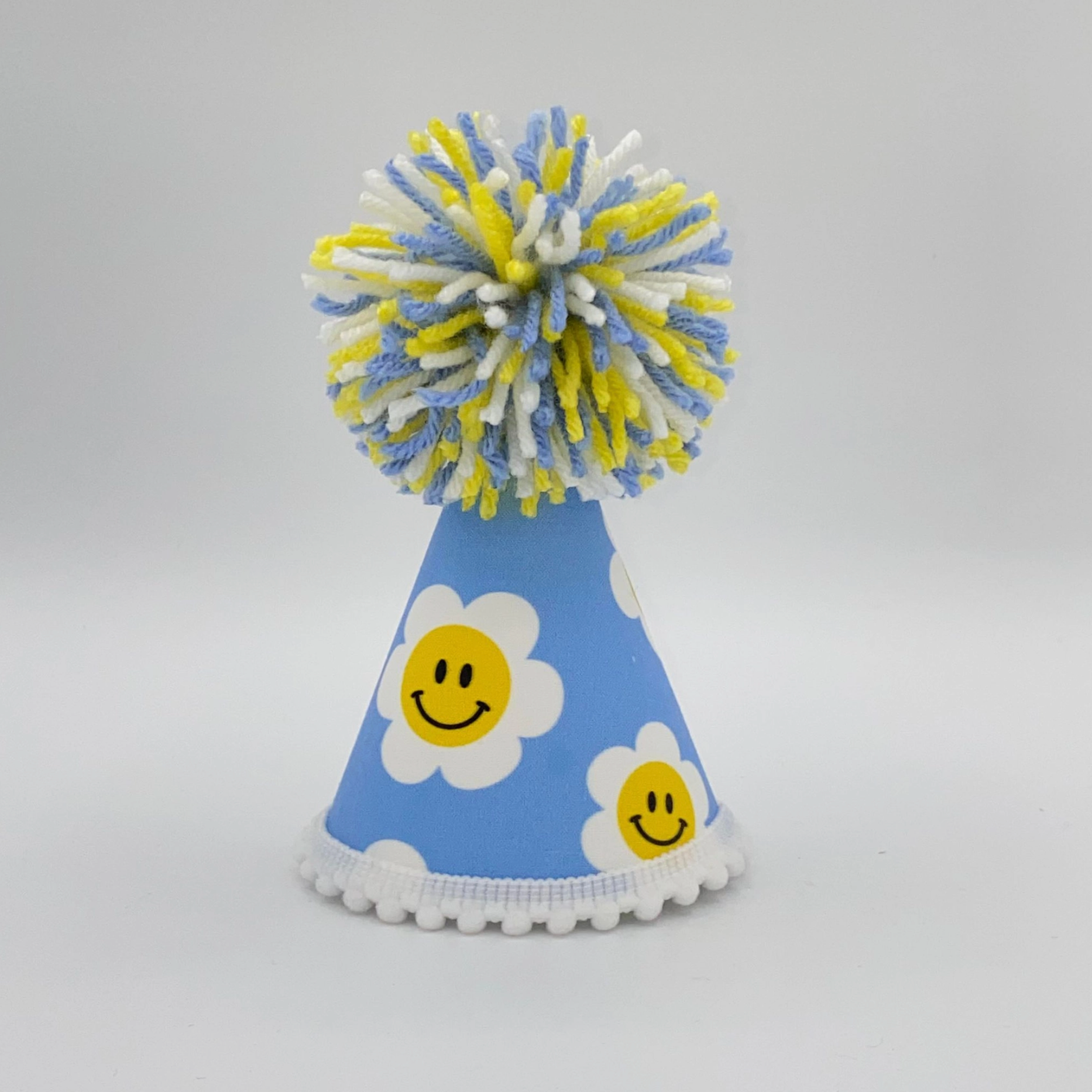 A blue pet party hat with a smiley face daisy design and a yellow pom pom on top.