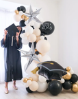 balloon garland with a graduate standing next to it, in black, white, cream and gold colors. starburst balloons are added in.
