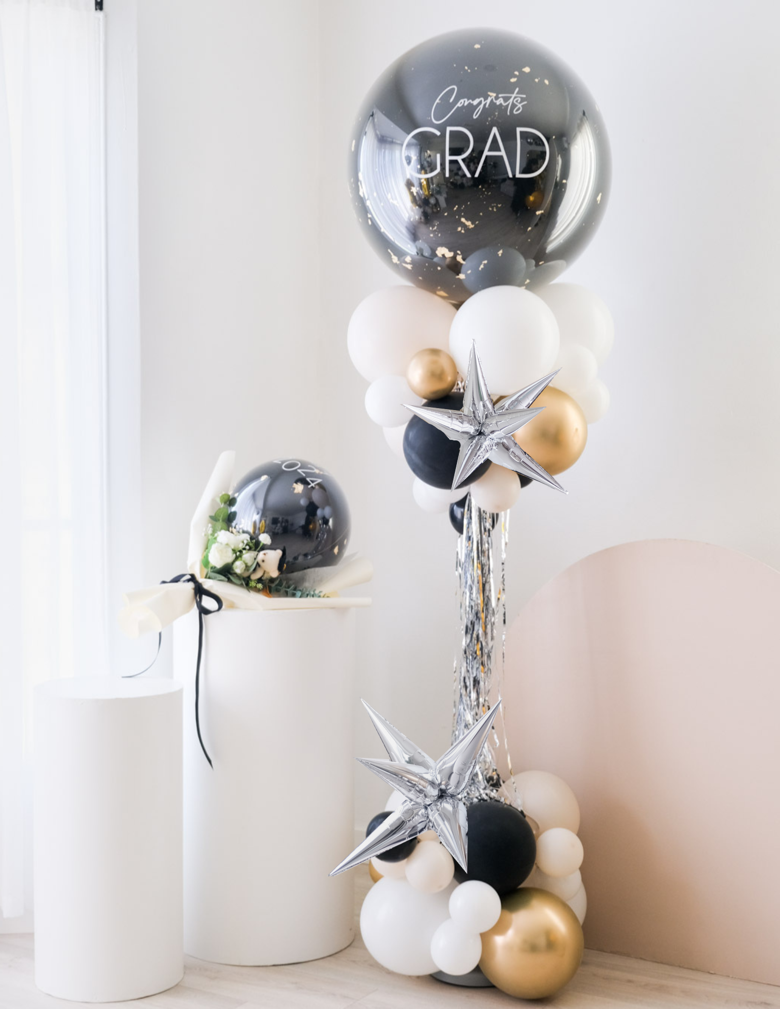A balloon tower with a combination of silver, black, white, and gold balloons, with  silver starburst balloons, with the top balloon saying 'Congrats GRAD'.