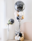 A balloon tower with a combination of silver, black, white, and gold balloons, with  silver starburst balloons, with the top balloon saying 'Congrats GRAD'.