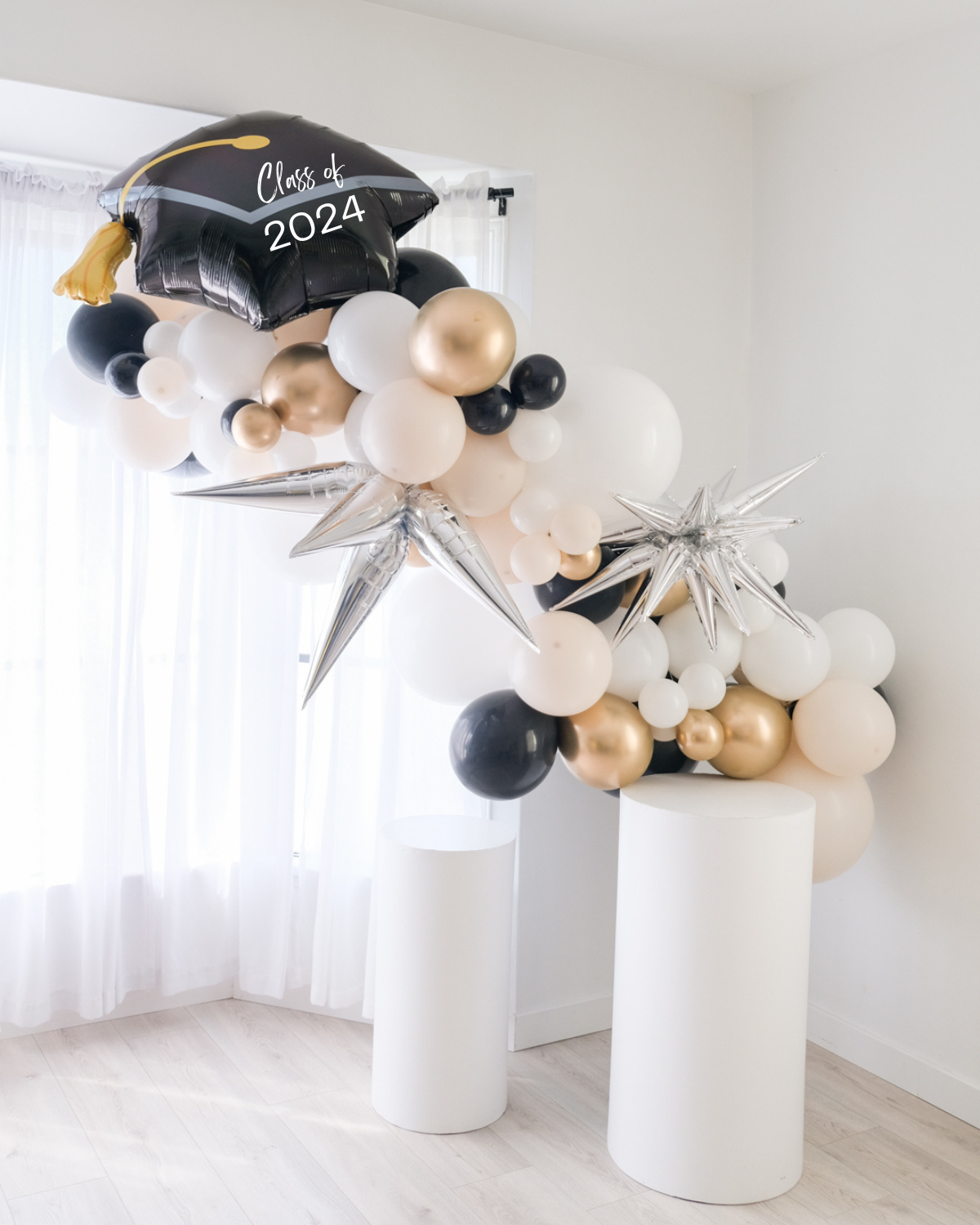 A balloon garland featuring a graduation cap design, with a mix of gold, silver, and white balloons, displayed indoors.