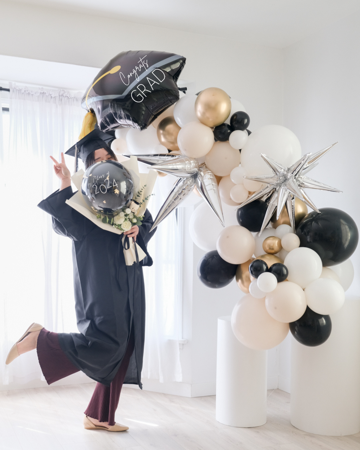 A balloon garland featuring a graduation cap design, with a mix of gold, silver, and white balloons, displayed indoors. A grad student wearing a gown and holding a balloon bouquet.