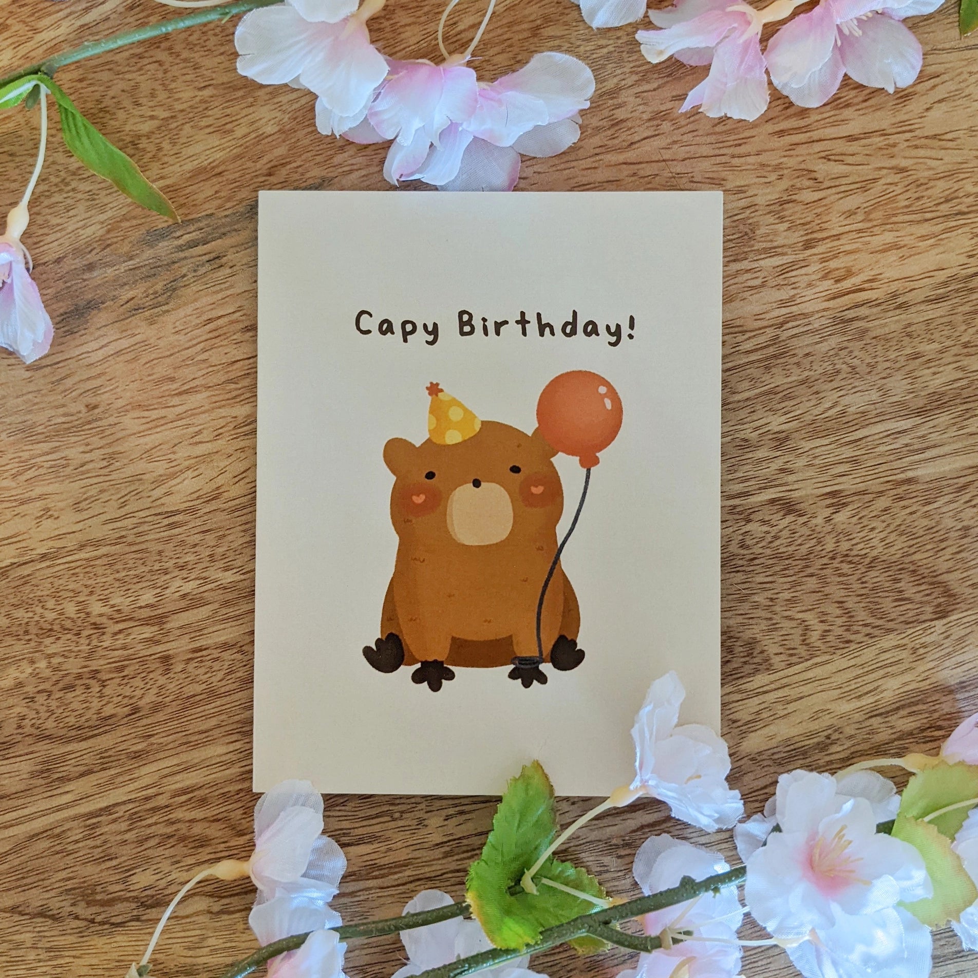 A birthday card with an illustration of a capybara holding a balloon, with the text 'Capy Birthday!' written on it. The card is placed on a wooden surface with artificial flowers around it.