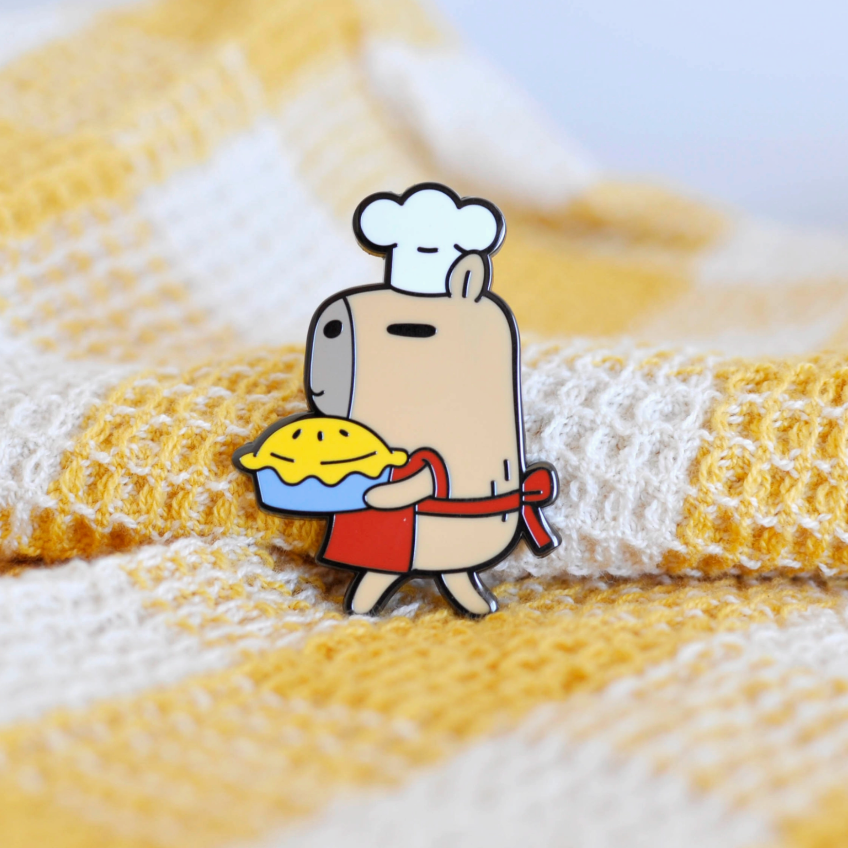 A black nickel plated enamel pin featuring a cartoon capybara character with a chef's outfit and a pie.