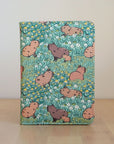 A passport wallet with a green background featuring a pattern of capybaras in various shades of pink and beige, with yellow floral accents.