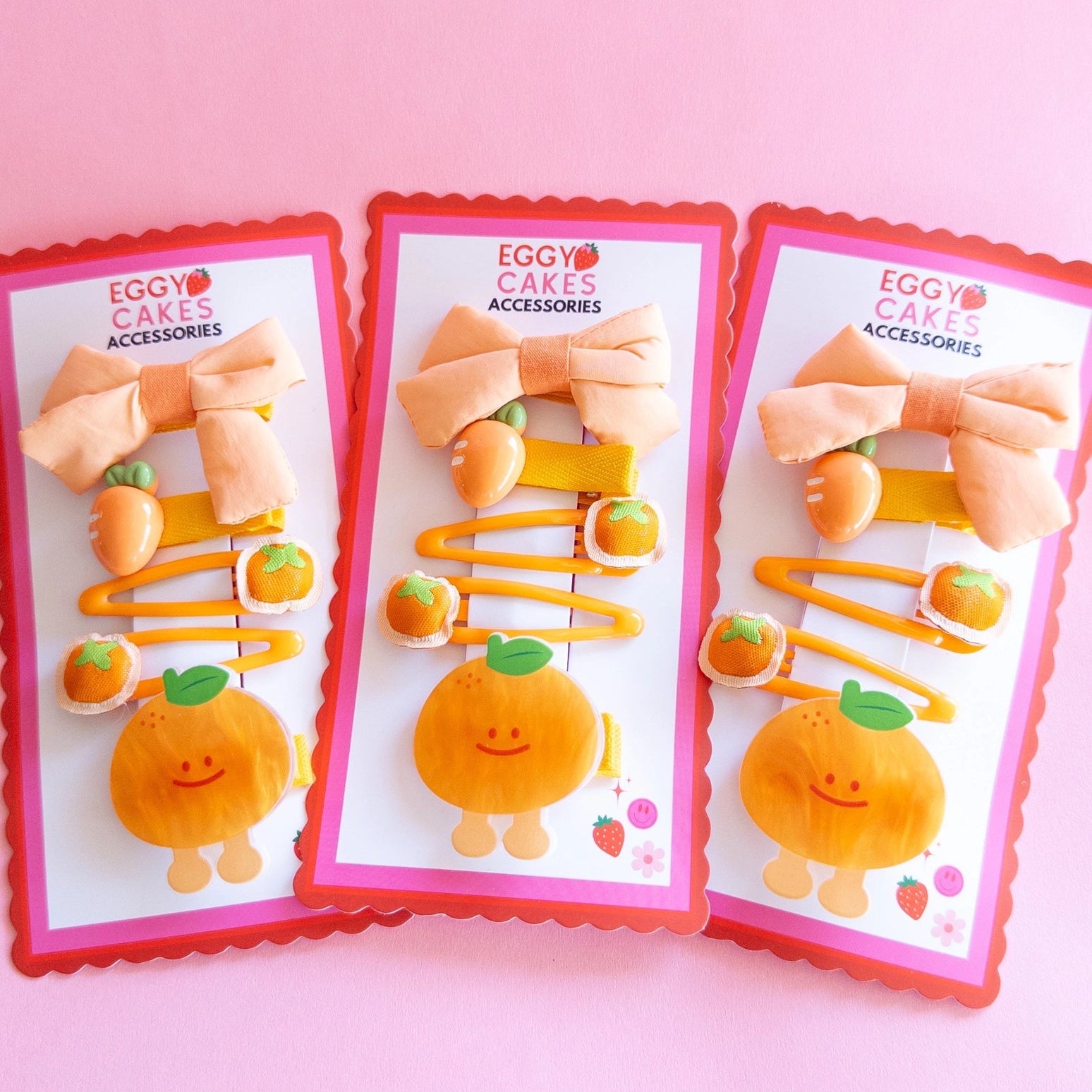 Three packaged sets of cute orange-themed hair accessories on a pink background.