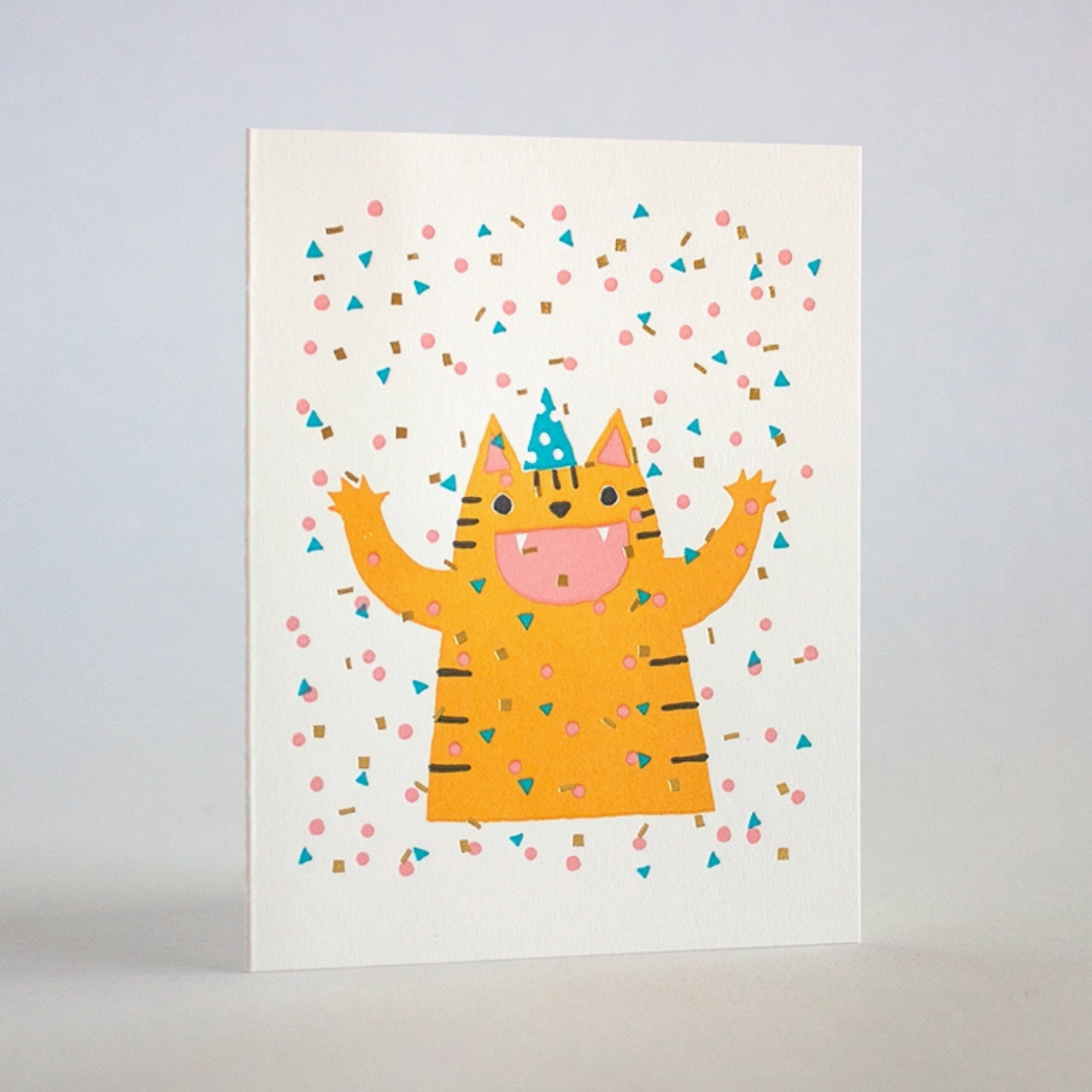 Greeting card with a cartoon illustration of a smiling cat wearing a party hat, surrounded by multicolored confetti on a white background. The card has gold foil details and a blank inside for a message, with a matching envelope.
