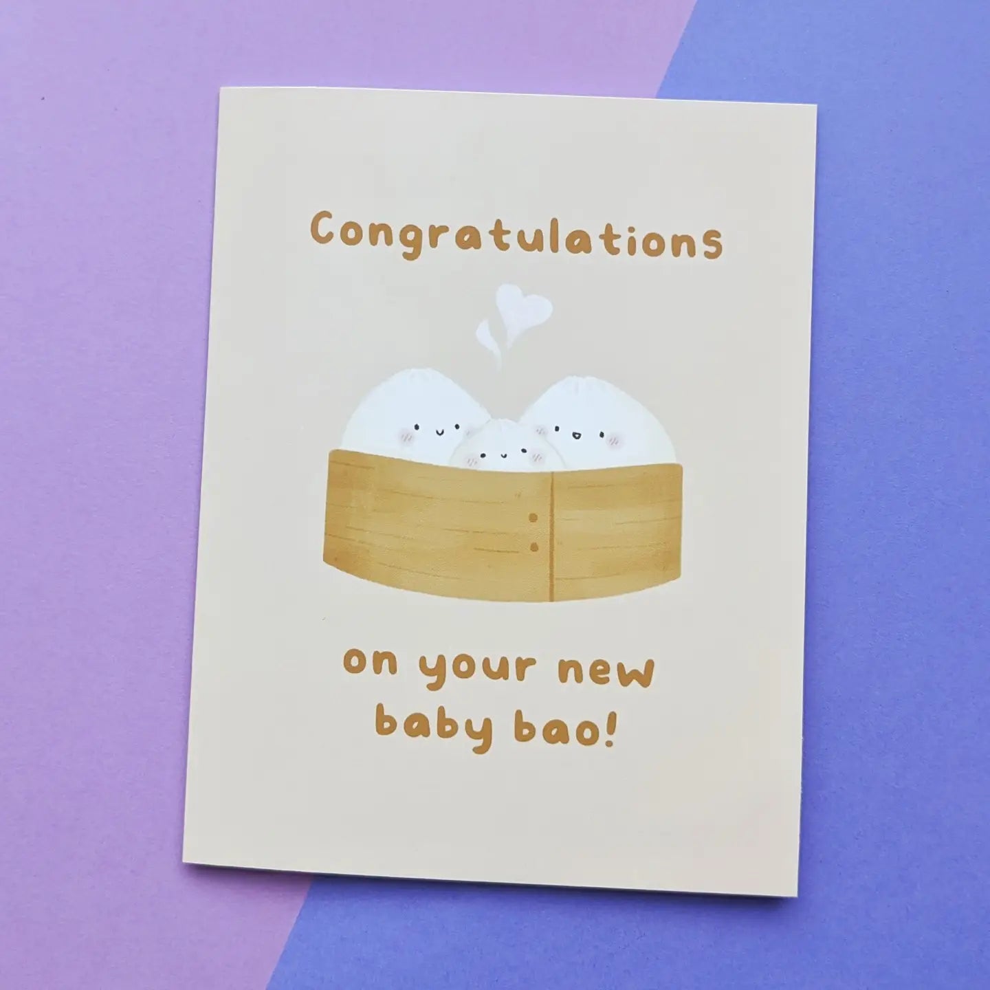 A congratulations card with a cute illustration of two white baby baos in a wooden steamer, with the text 'Congratulations on your new baby bao!' in brown, shown on a split purple and blue background.
