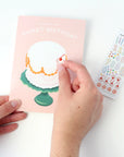 Hand sticking a sticker of a cake onto a 'Sweet Birthday' card with a second card featuring numbers and stickers in the background.