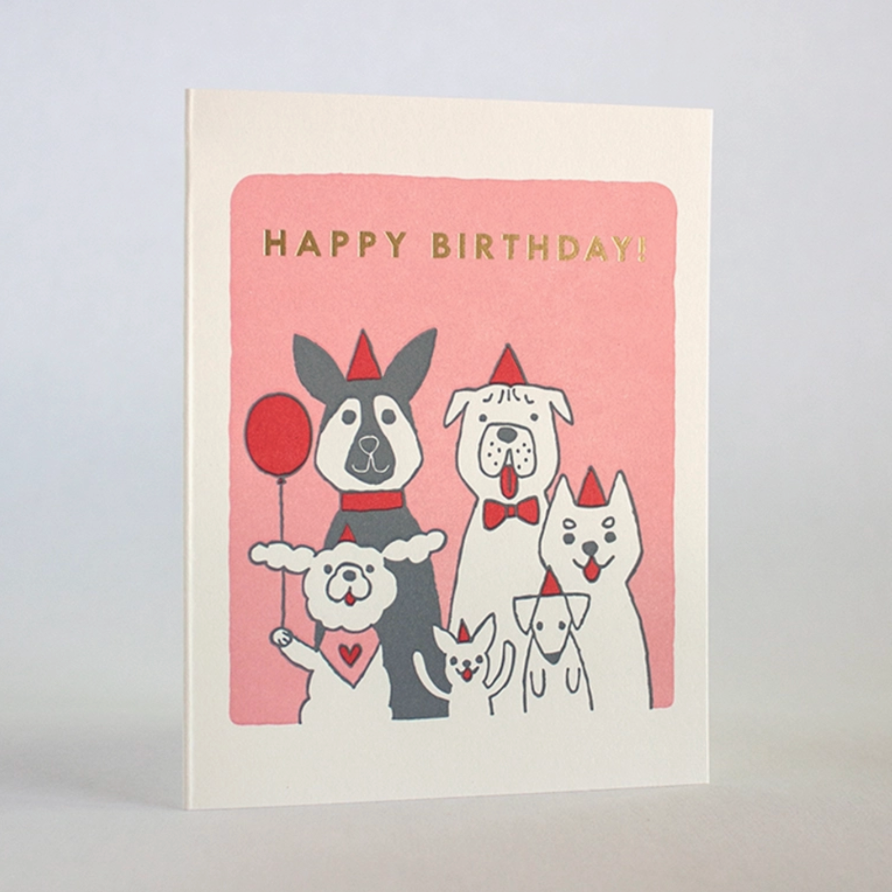 A greeting card with an illustration of various dogs in party hats and costumes on a pink background, with 'Happy Birthday!' text in gold foil details.