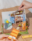 A miniature DIY dollhouse with a garden house theme, featuring various furniture, potted plants, and gardening tools. A hand using tweezers to place a box of flowers inside the house