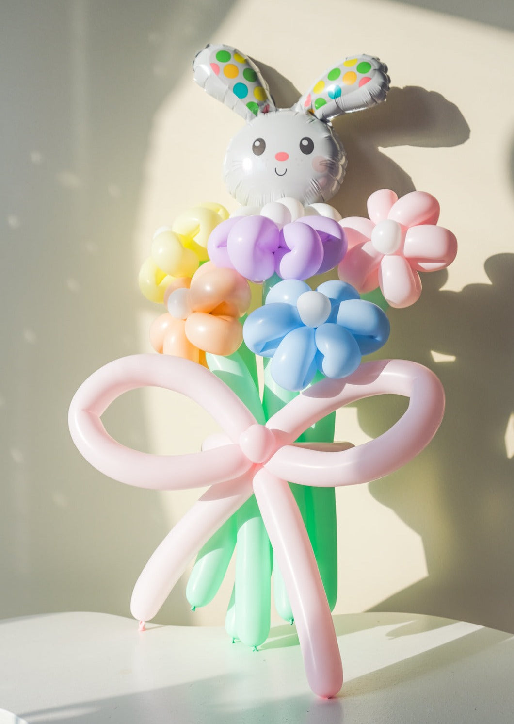 A balloon bouquet featuring a cute, cartoon-style bunny balloon with a bow, surrounded by various round balloons in pastel colors.
