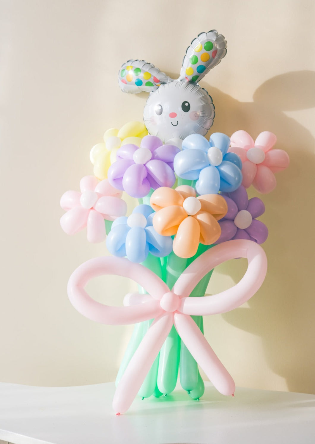 A balloon bouquet featuring a cute, cartoon-style bunny balloon with a bow, surrounded by various round balloons in pastel colors.
