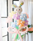 A balloon bouquet featuring a cute, cartoon-style bunny balloon with a bow, surrounded by various round balloons in pastel colors.