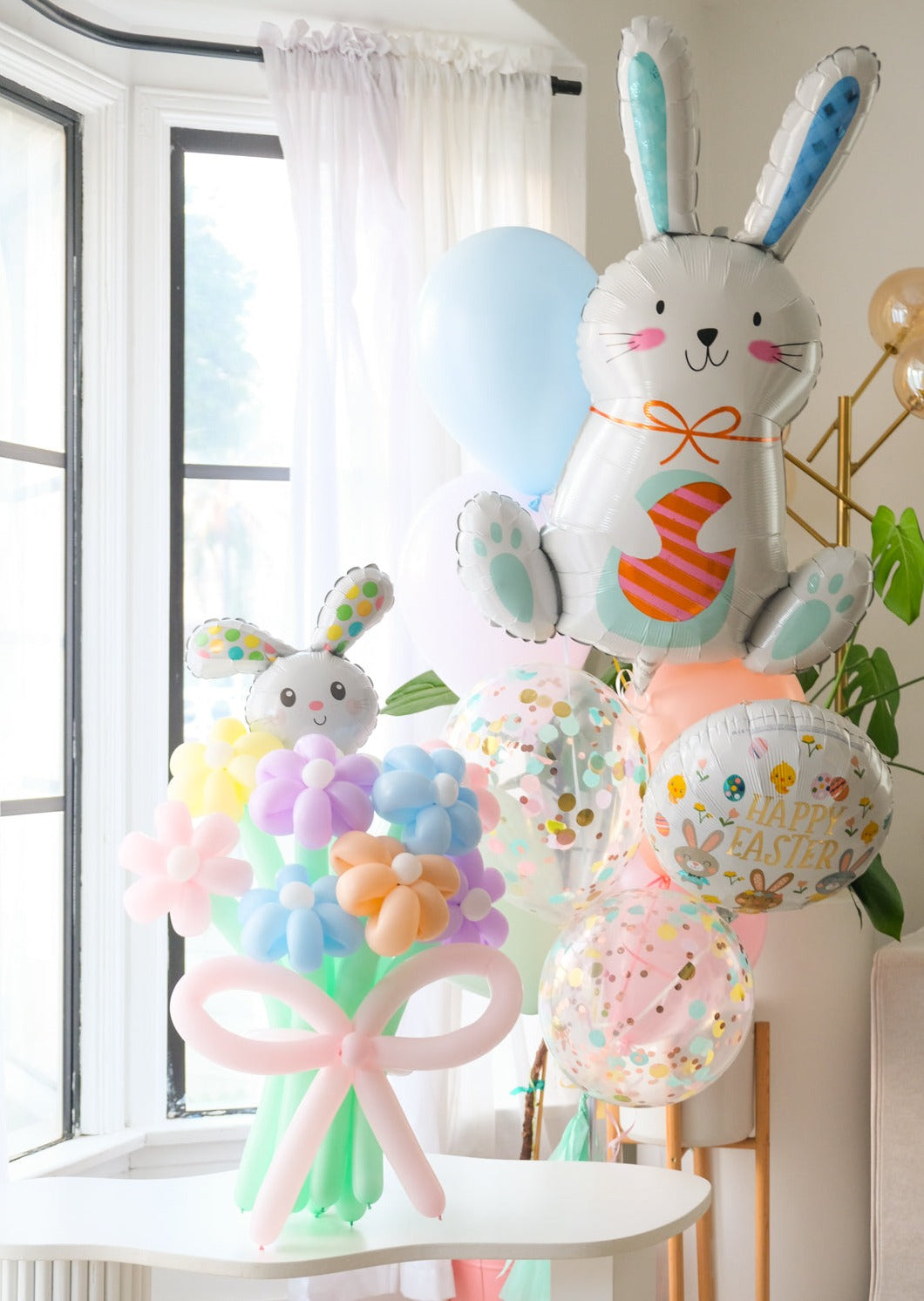 A balloon bouquet featuring a cute, cartoon-style bunny balloon with a bow, surrounded by various round balloons in pastel colors. Another helium bouquet with bunny and easter themed balloons behind it.
