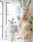 A balloon bouquet featuring a cute, cartoon-style bunny balloon with a bow, surrounded by various round balloons in pastel colors. Another helium bouquet with bunny and easter themed balloons behind it.