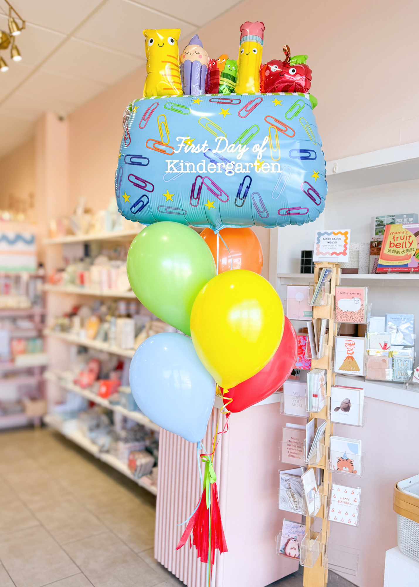 A balloon arrangement featuring a pencil pouch balloon, with colorful latex balloons, a handmade tassel, and a personalized grade tag, displayed in a store setting.