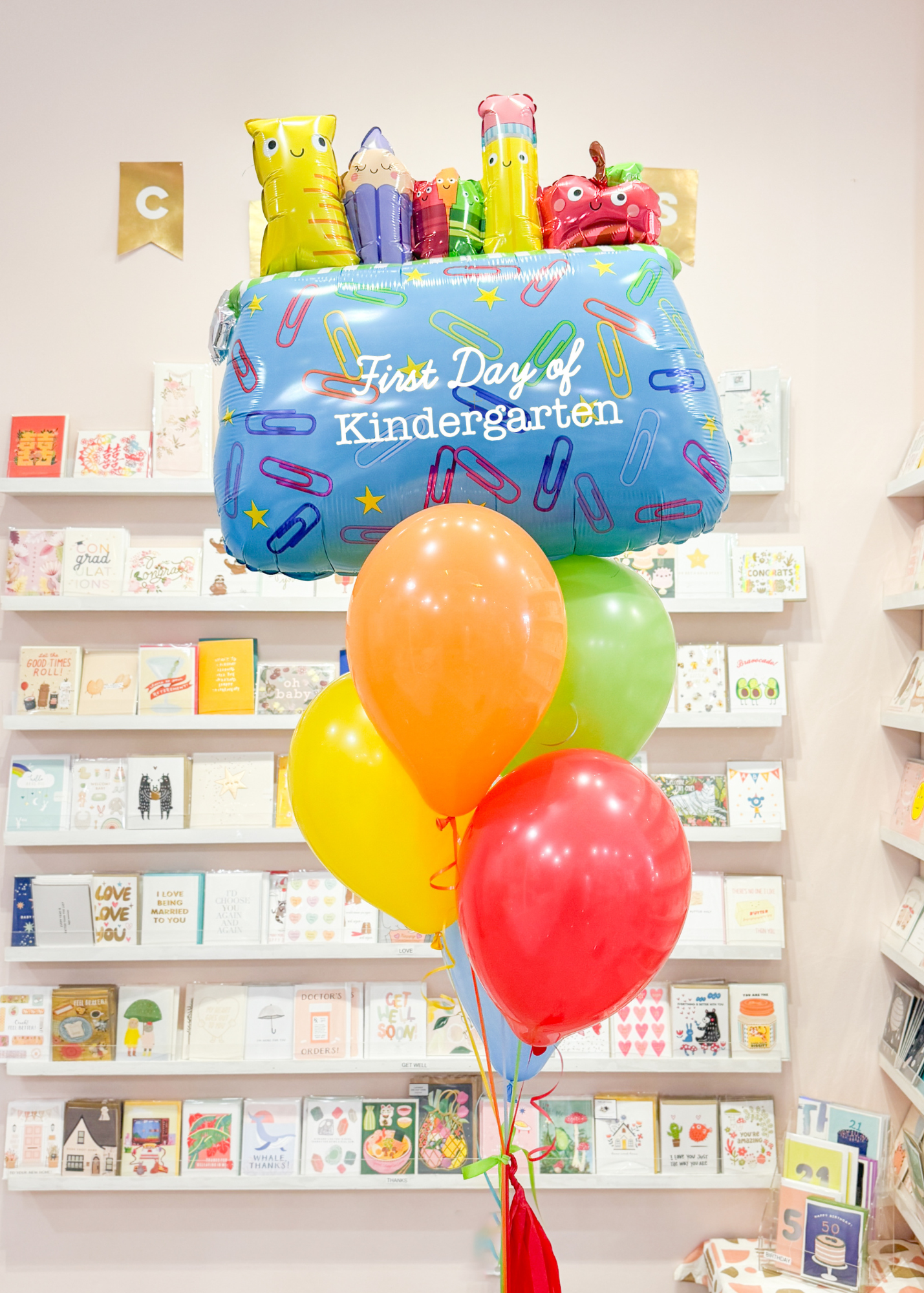 A balloon arrangement featuring a pencil pouch balloon, with colorful latex balloons, a handmade tassel, and a personalized grade tag, displayed in a store setting with card shelves