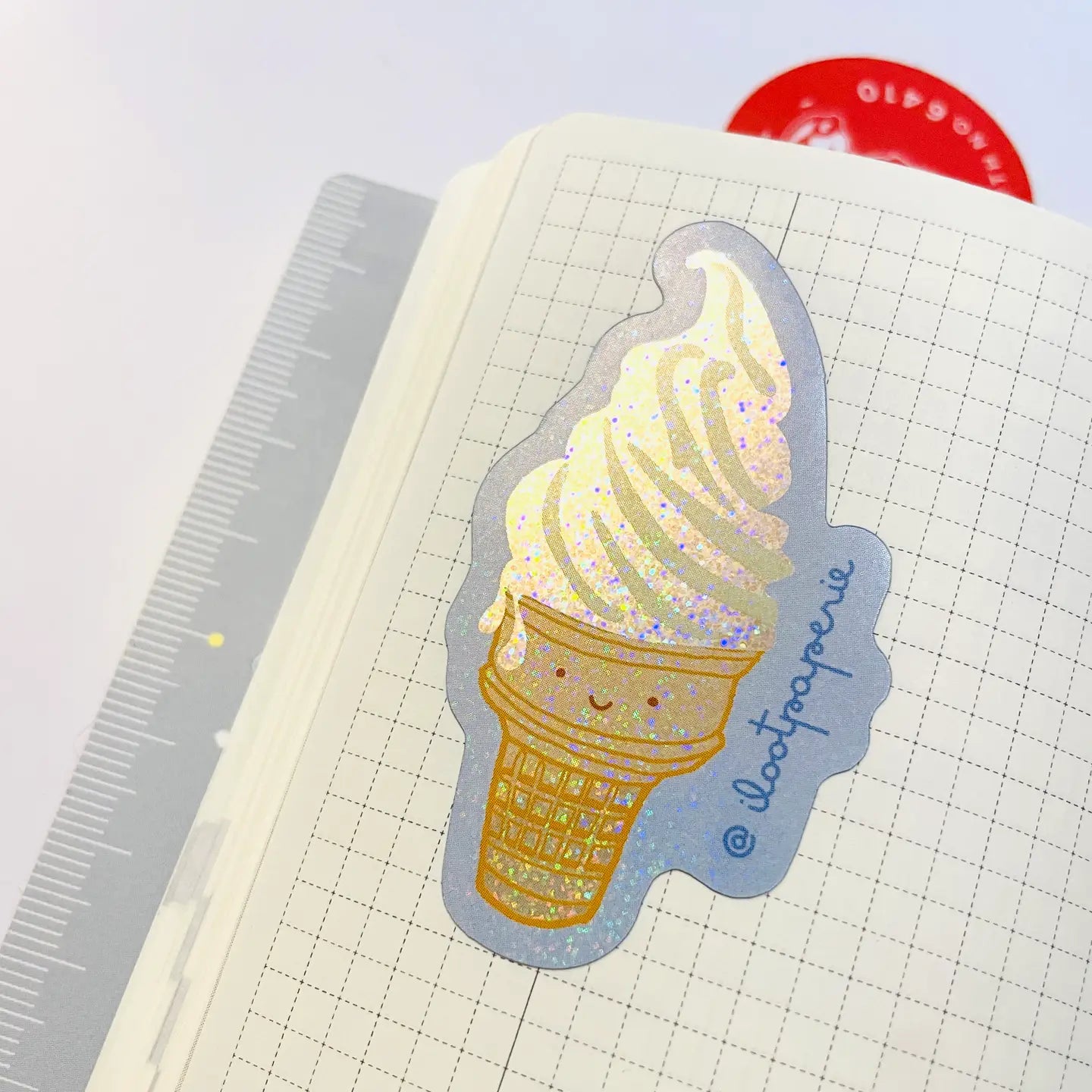 Sticker of a smiling ice cream cone on a notebook with a ruler and red object in the background.