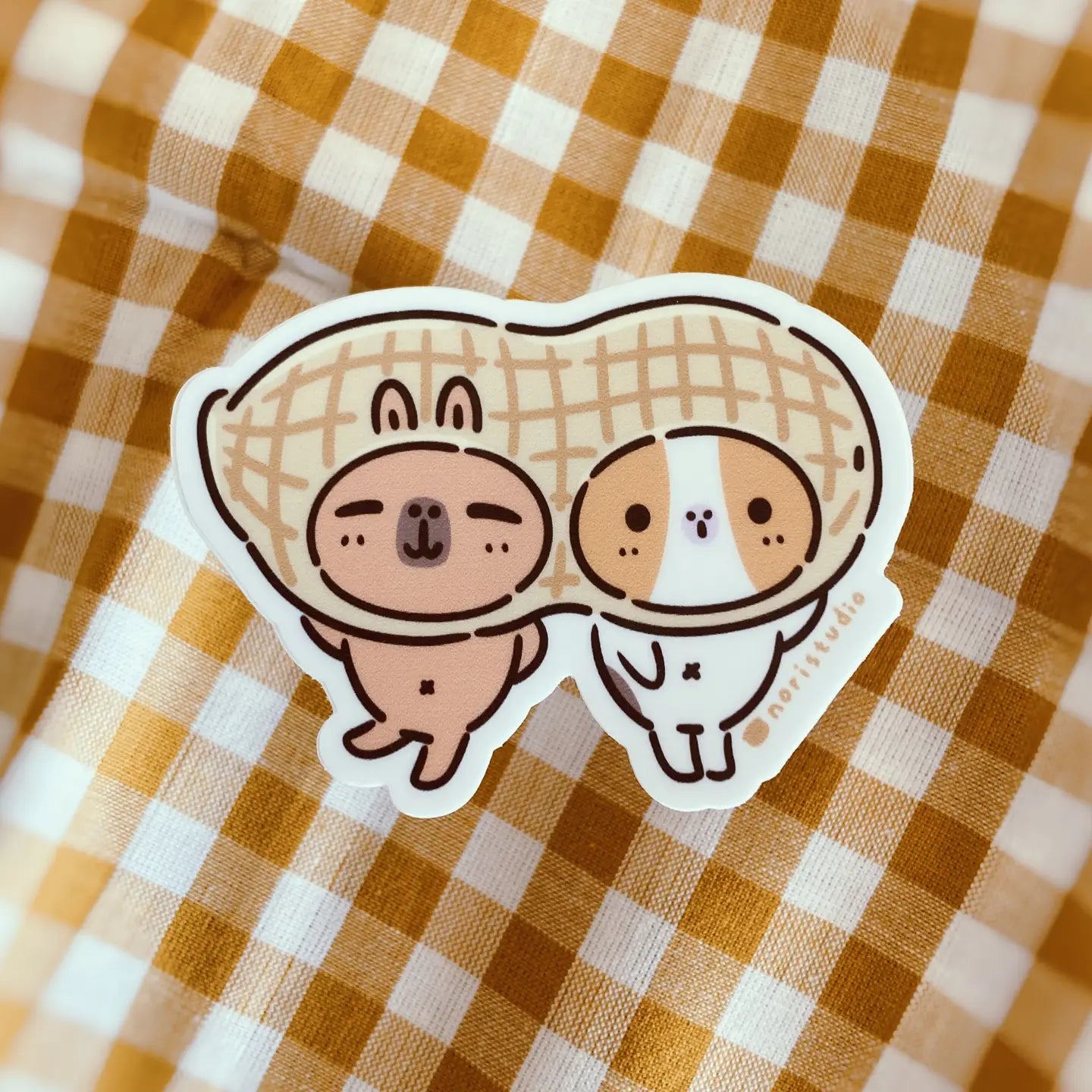A vinyl sticker featuring cartoon versions of a guinea pig and capybara, designed to look like peanuts, placed on a checkered background.