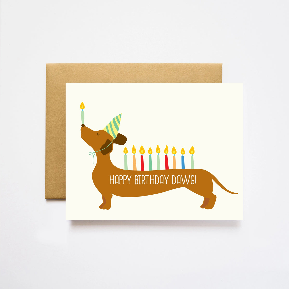 A birthday greeting card for a dachshund, featuring a cartoon brown dachshund wearing a green striped party hat and balancing a single candle on its nose while holding a row of colorful candles on its back. The card reads 'HAPPY BIRTHDAY DAWGI' in white text.