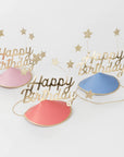 Three 'Happy Birthday' hats in pink, blue, and red with gold stars on a light gray background.