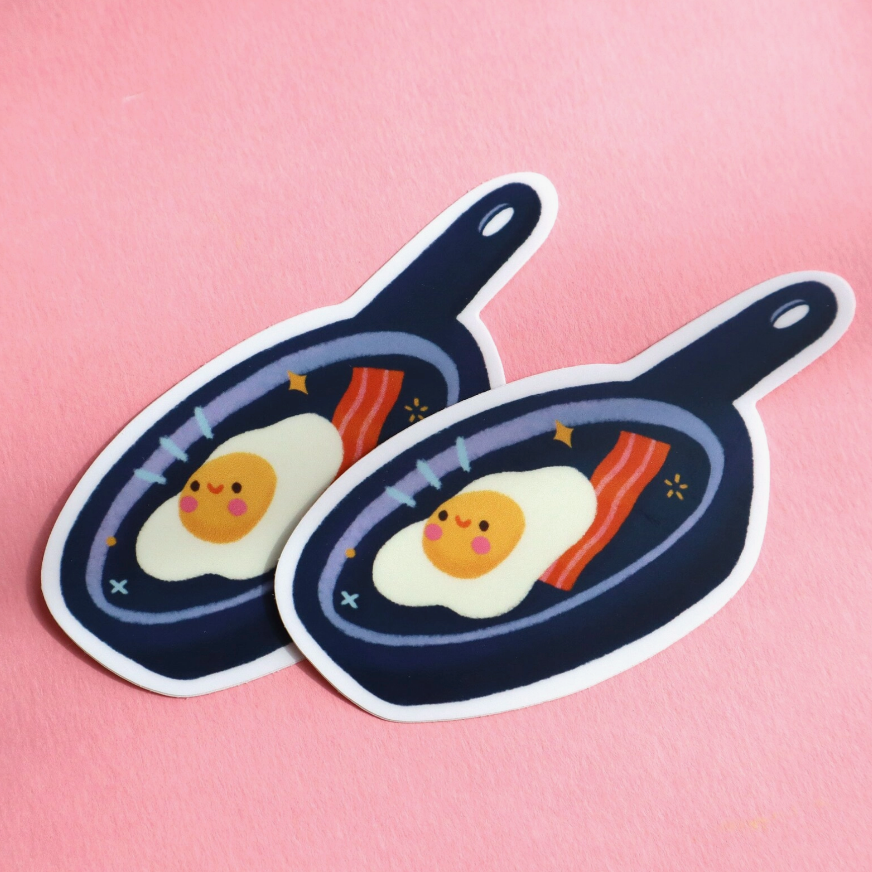 Two stickers depicting a cartoon of an egg and a piece of bacon frying in a pan, with a pink background.