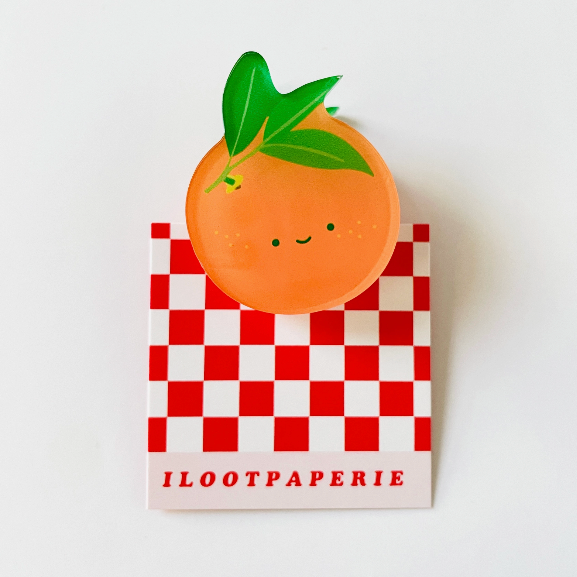 A clear acrylic clip designed to look like an orange with a smiling face, placed on a red and white checkered surface.