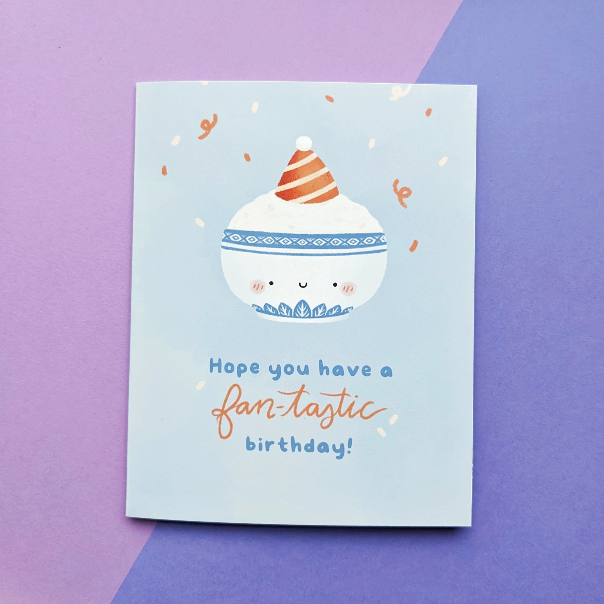 A birthday card with a graphic of a bowl of rice wearing a party hat, accompanied by text 'Hope you have a fan-tastic birthday!' on a light blue background. A purple envelope is visible behind the card.