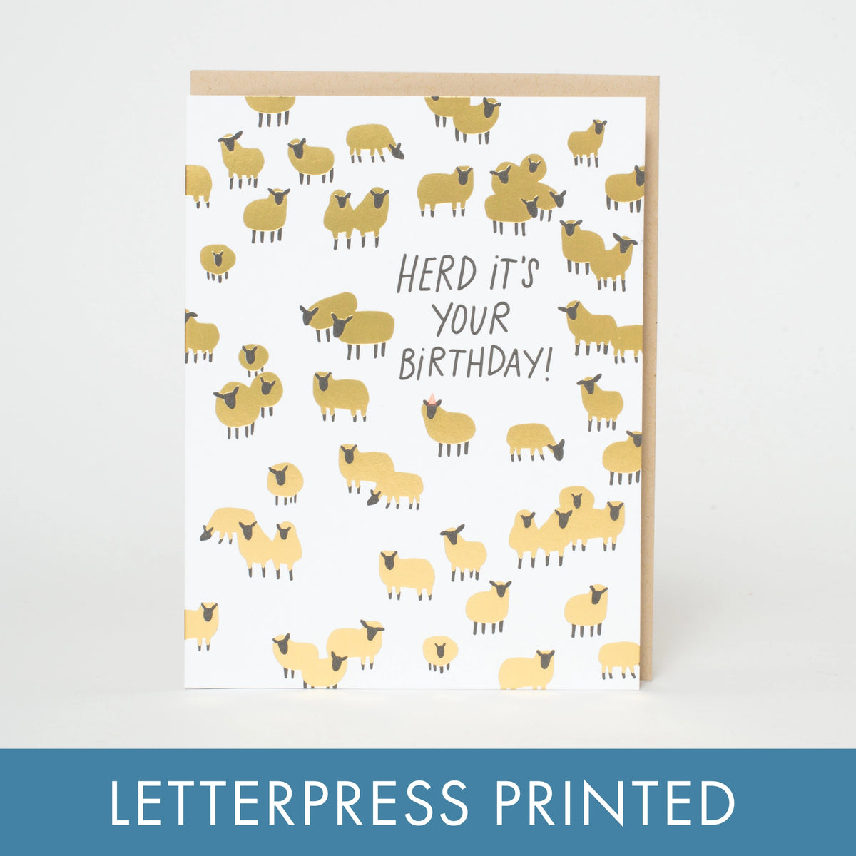 A white greeting card with a repeating pattern of small yellow sheep and the text 'HERD IT'S YOUR BIRTHDAY!' in black, shown with a kraft envelope behind it on a white background. A blue banner at the bottom reads 'LETTERPRESS PRINTED'.