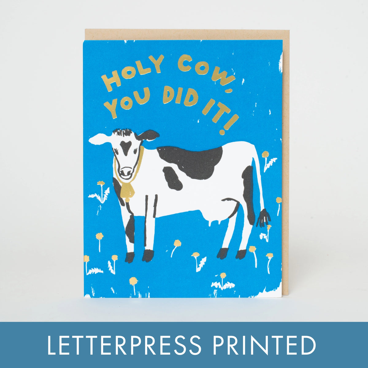 A letterpress greeting card featuring a blue background with the text 'HOLY COW, YOU DID IT!' in yellow above a black-and-white cow illustration. A kraft envelope is partially visible behind the card, and a blue banner at the bottom reads 'LETTERPRESS PRINTED'.