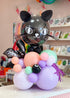A decorative balloon arrangement shaped like a black cat head on a cluster of pastel balloons, including lavender, peach, mint green, pink, and purple, photographed in a retail or party-supplies setting.