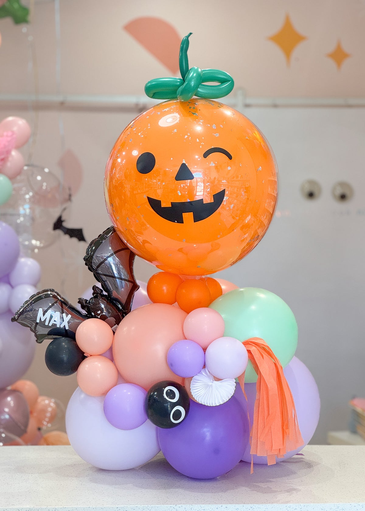 A Halloween-themed balloon bouquet featuring a large orange jack-o'-lantern balloon with a green stem, on top of a stack of balloons in pastel colors along with black bat balloon featuring a personalized name