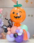A Halloween-themed balloon bouquet featuring a large orange jack-o'-lantern balloon with a green stem, on top of a stack of balloons in pastel colors along with black bat balloon featuring a personalized name