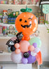 A Halloween-themed balloon bouquet featuring a large orange jack-o'-lantern balloon with a green stem, on top of a stack of balloons in pastel colors along with black bat balloon
