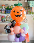 A Halloween-themed balloon bouquet featuring a large orange jack-o'-lantern balloon with a green stem, on top of a stack of balloons in pastel colors along with black bat balloon