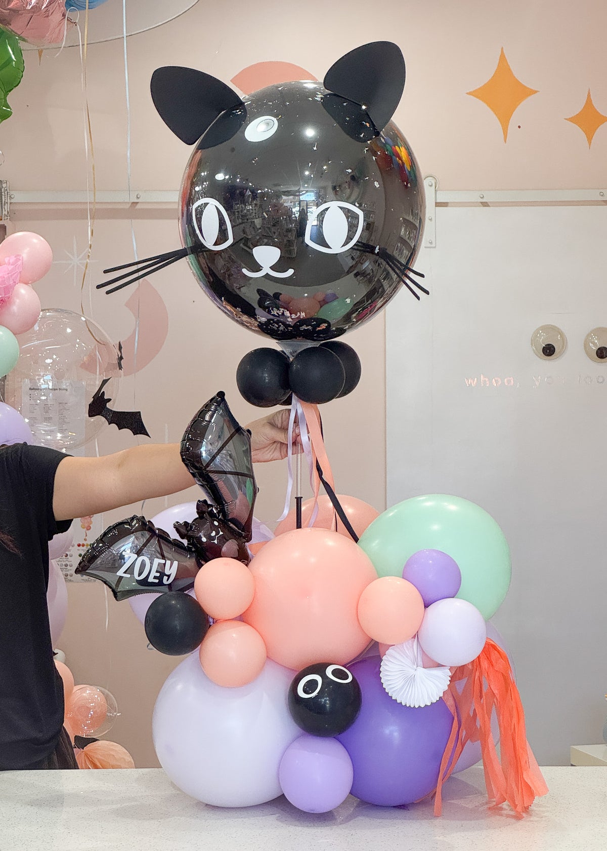 A decorative balloon arrangement featuring a large glossy black cat-shaped balloon with white eyes and ears on wand, held above pastel pink, purple, mint green, and lavender balloons, with orange tassel accents. 