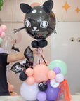A decorative balloon arrangement featuring a large glossy black cat-shaped balloon with white eyes and ears on wand, held above pastel pink, purple, mint green, and lavender balloons, with orange tassel accents. 
