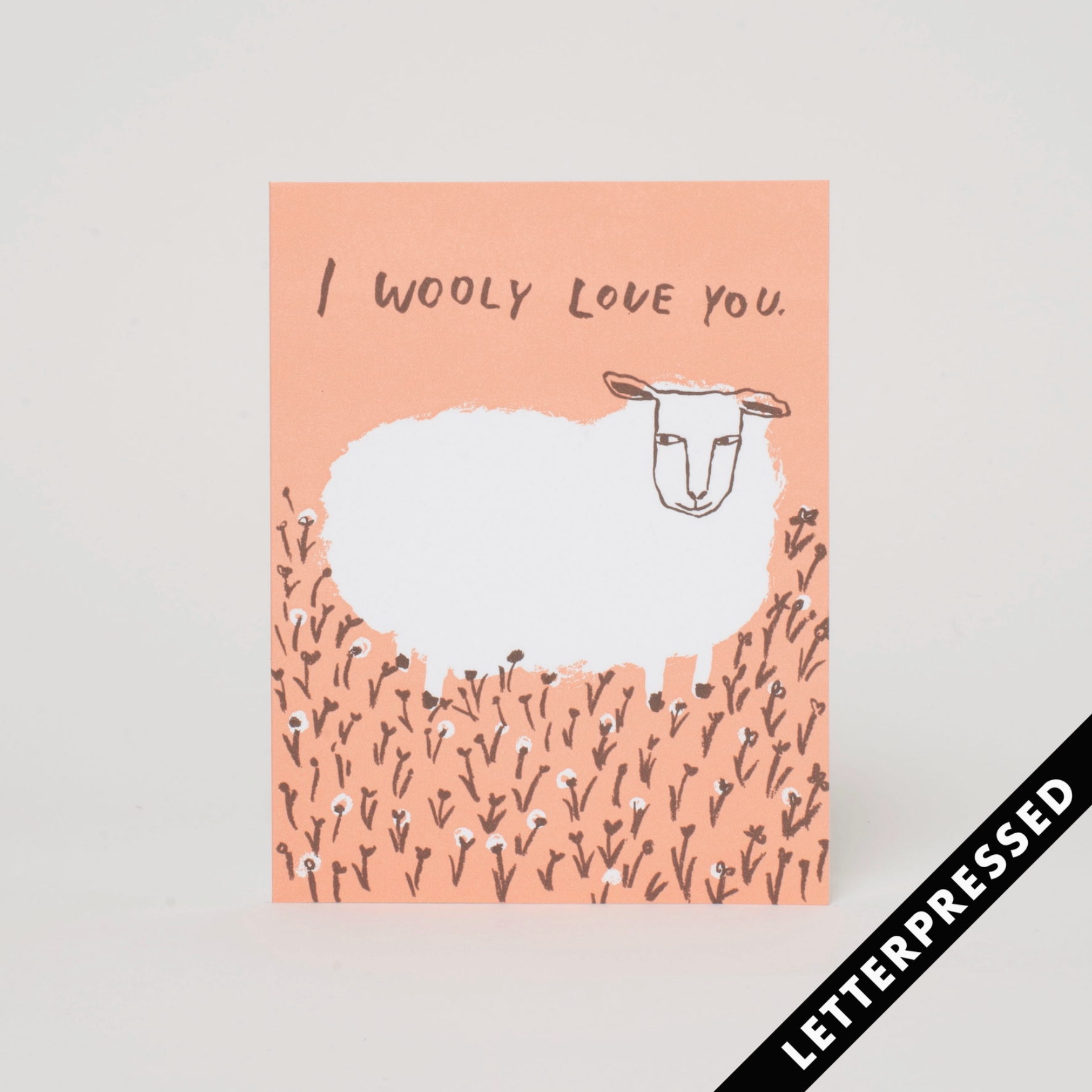 A greeting card with a sheep in a field and peach background, text reads "I wooly love you"