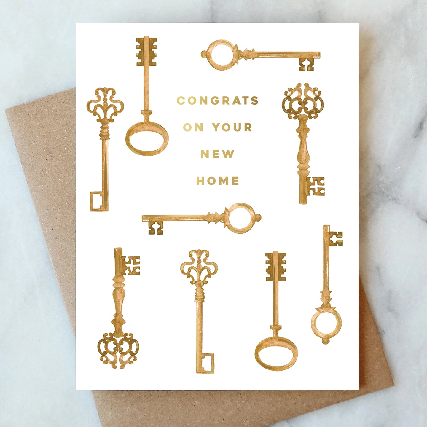 A white greeting card with gold foil decorative keys and the text 'CONGRATS ON YOUR NEW HOME,' placed on top of a brown kraft envelope over a light marble surface.