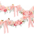 A section of a floral garland with pink and peach tissue paper flowers, pink and gold ribbons, and green artificial foliage.