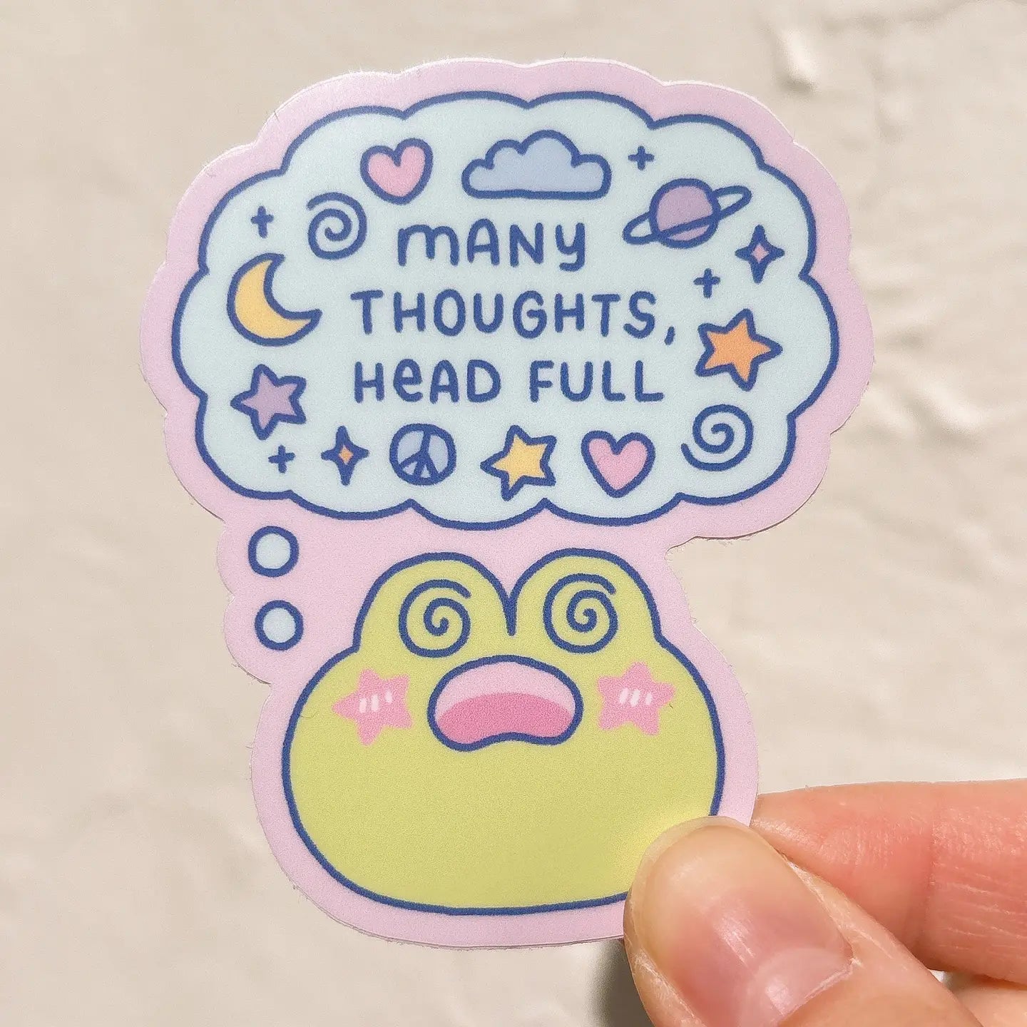 Sticker with text 'Many thoughts, head full' and various symbols on a light background above a frog that is dizzy