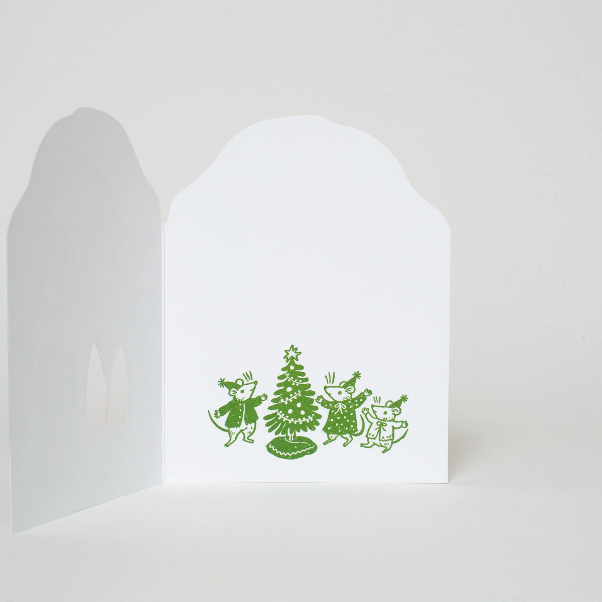 A white greeting card shown open against a light gray background, featuring a green line-art illustration of three children dancing around a Christmas tree.